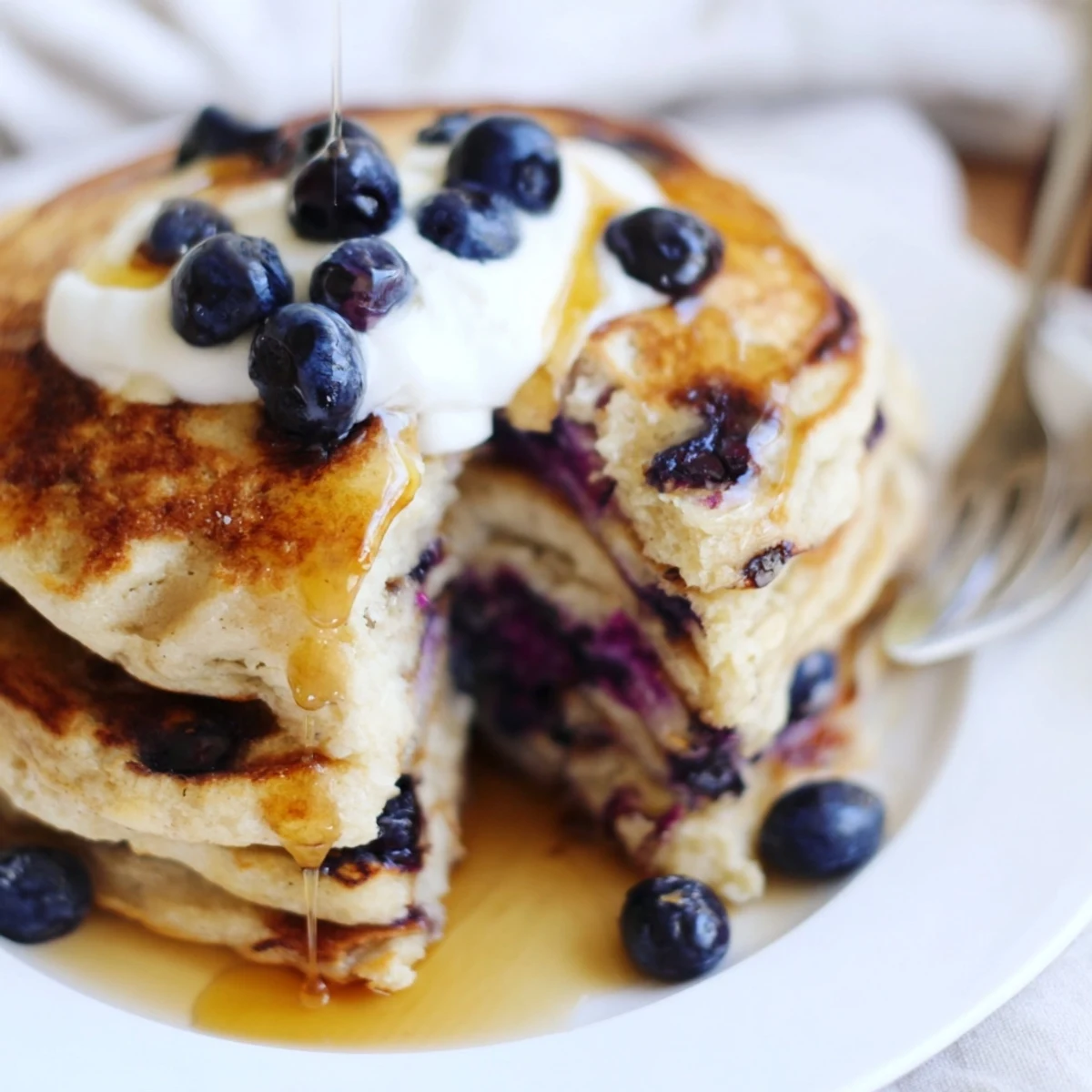 Golden brown Greek yogurt blueberry pancakes stacked high on a white plate with sweet maple syrup glaze