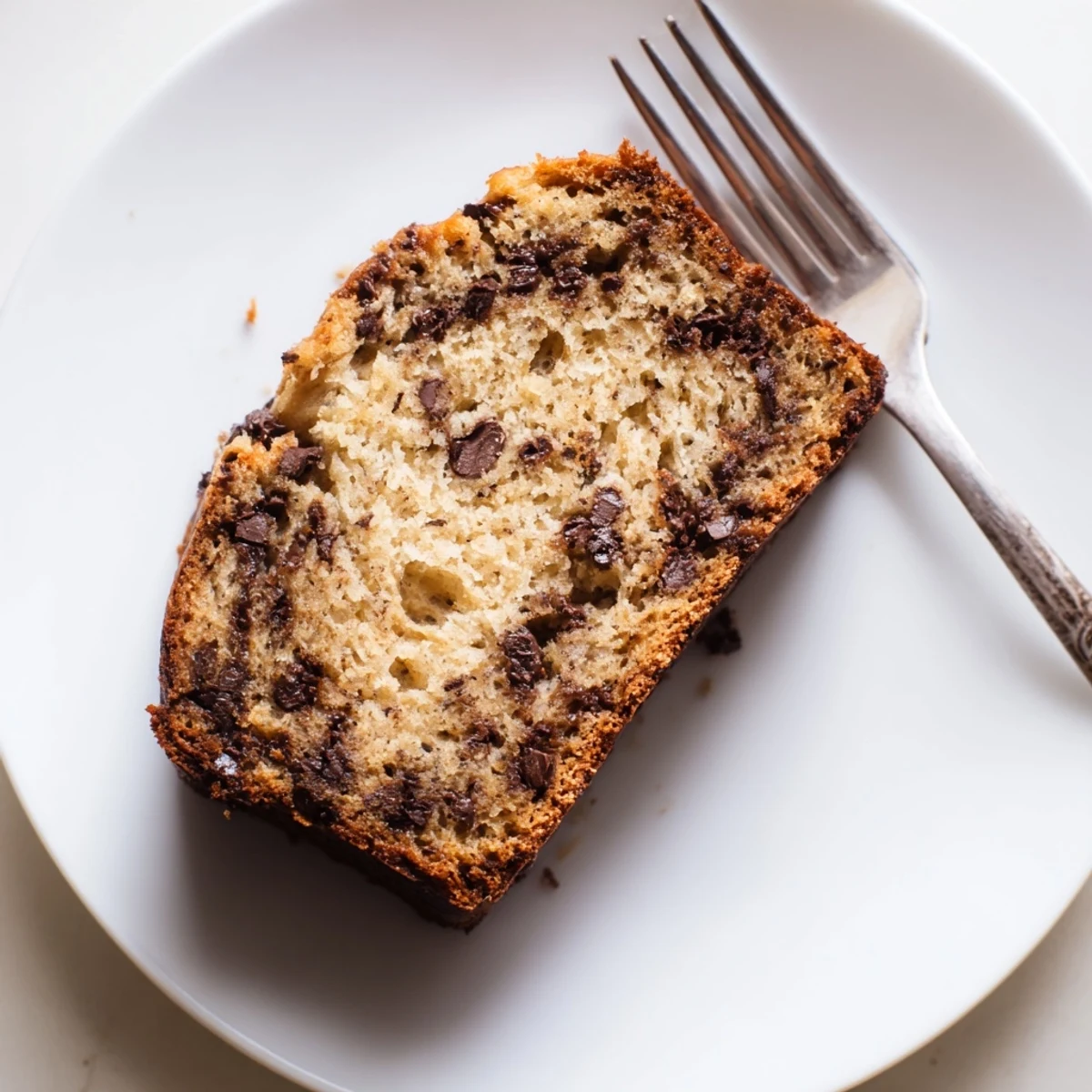 Slice of moist chocolate chip banana bread revealing gooey chips on a white plate